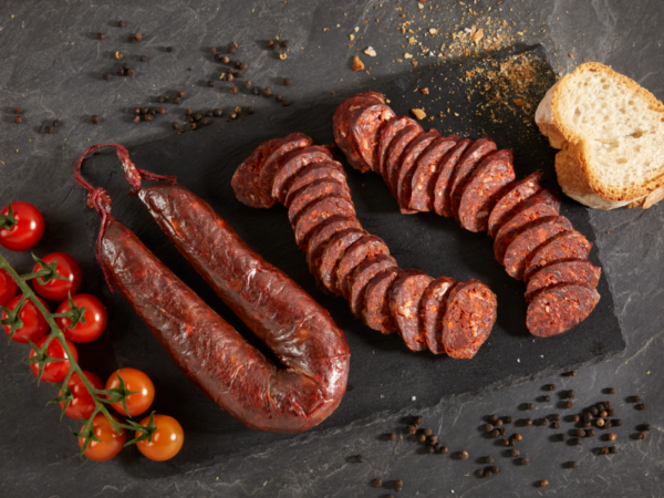 CHORIZO VACUNO MAYOR ASTURIANO Sobre un fondo oscuro hay en el centro dos chorizos (uno sin cortar y otro troceado). Rodeando las salchicas hay tomates y un trozo de pan, a izquierda y derecha respectivamente