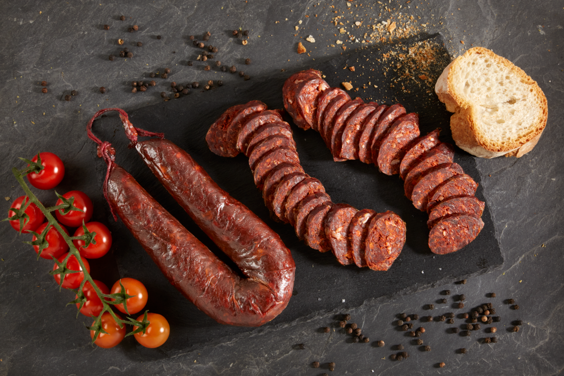 CHORIZO VACUNO MAYOR ASTURIANO Sobre un fondo oscuro hay en el centro dos chorizos (uno sin cortar y otro troceado). Rodeando las salchicas hay tomates y un trozo de pan, a izquierda y derecha respectivamente